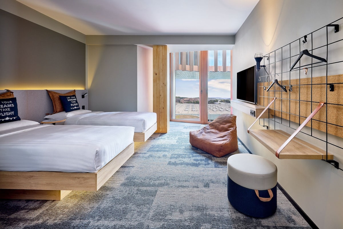 A modern hotel room with two beds, a large window, a TV mounted on a wall, and a wooden bench.