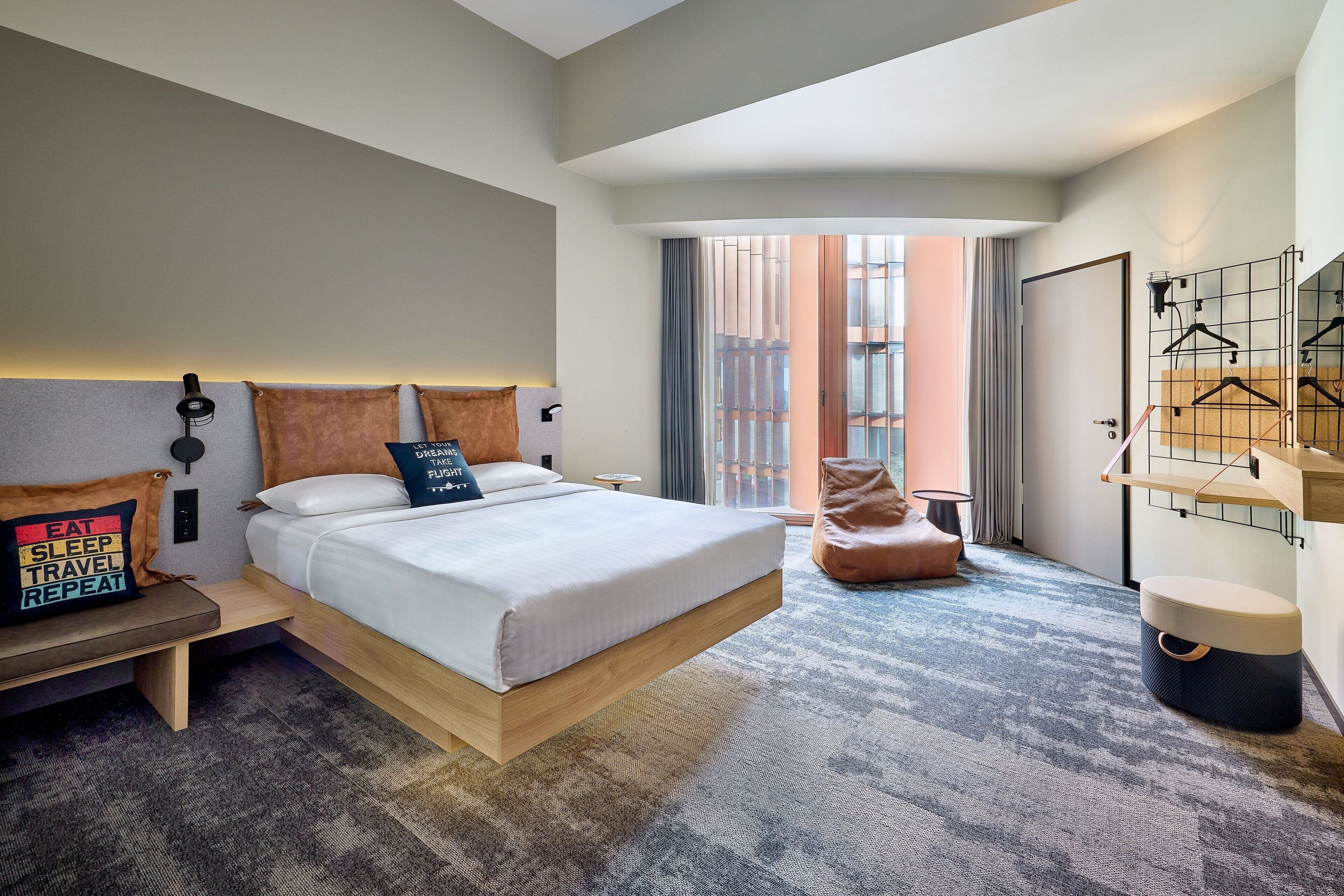 A modern hotel room with a large bed, bedside tables, a bench, a seating area, and a view of the city through large windows.