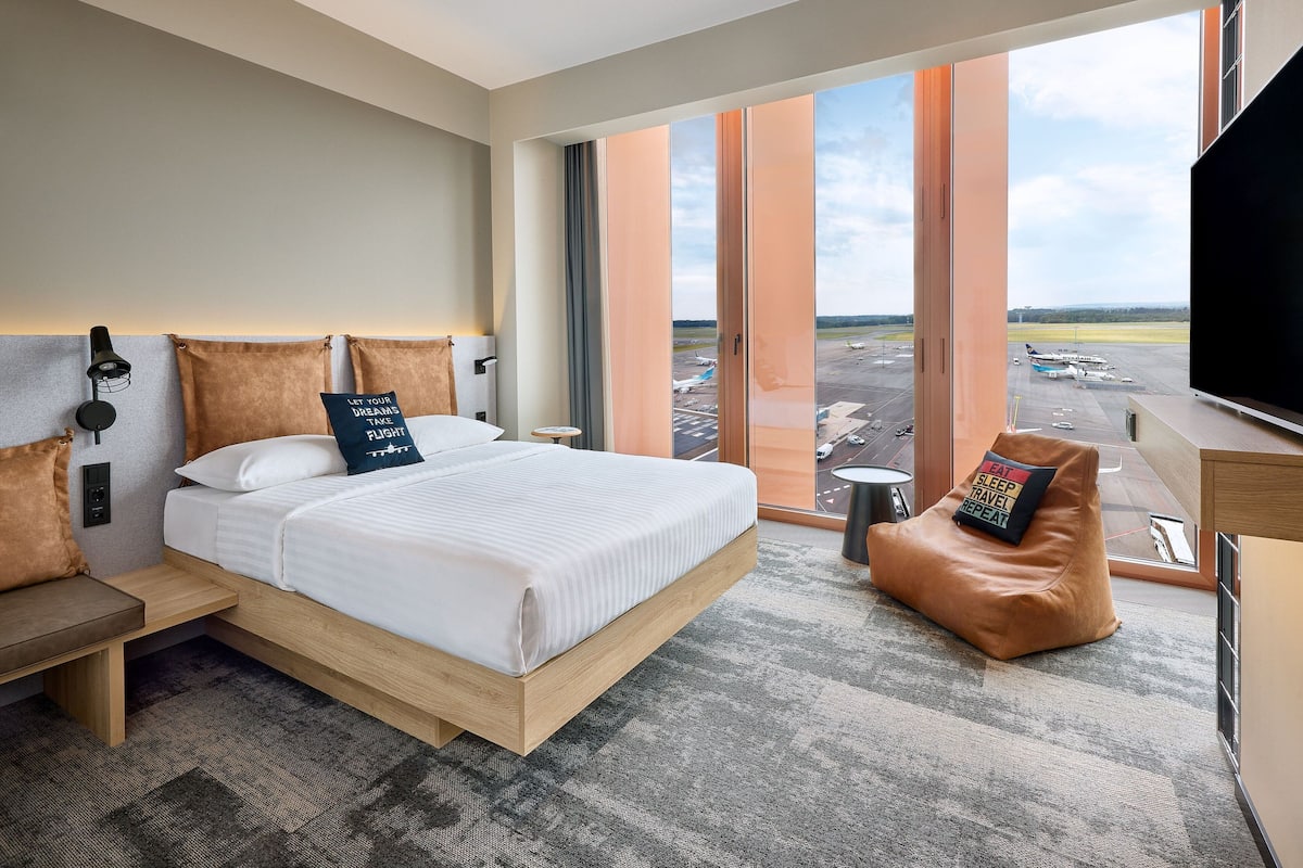 A modern hotel room with a large bed, a seating area, and a view of the airport.