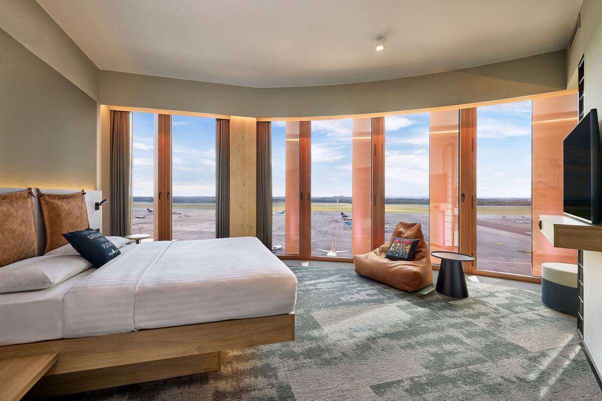 A modern hotel room with a large bed, a seating area, and a view of the airport.