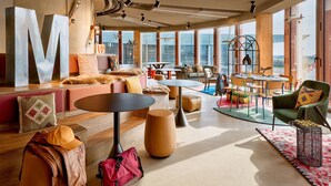 Een moderne lounge met een groot ‘M’ sculptuur, zachte zitplaatsen en een stadszicht door grote ramen.