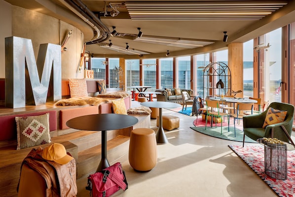 A modern lounge area with a large 'M' sculpture, comfortable seating, and a view of the city through large windows. - Moxy Luxembourg Airport (Sandweiler)