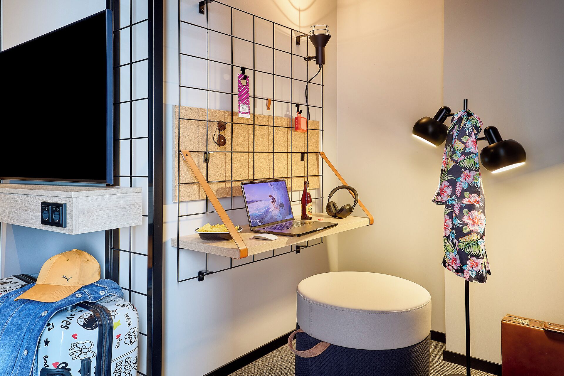 A modern hotel room with a TV, a laptop on a shelf, a round ottoman, and a wall-mounted shelf with various items.