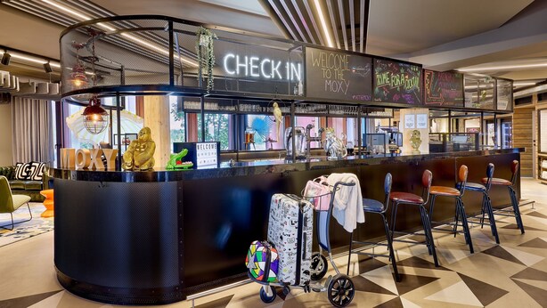 A check-in counter with a 'CHECK IN' sign, a 'WELCOME TO THE MOXY' sign, and a 'TIME FOR A COCONUT' sign. There are chairs and a luggage cart with suitcases and a backpack. - Moxy Luxembourg Airport (Sandweiler)