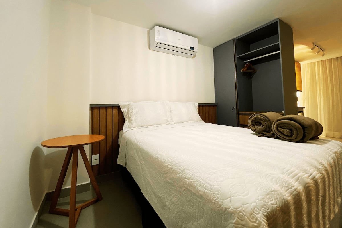Ein Hotelzimmer mit einem Bett, einem kleinen Holfnachtkästchen, einer Klimaanlage, einem Kleiderschrank und einem Badezimmer mit Duschvorhang.