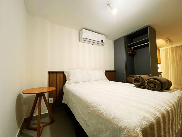 A hotel room with a bed, a small wooden side table, an air conditioning unit, a wardrobe, and a bathroom with a shower curtain.