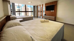 A hotel room with two beds, a small round table, a TV mounted on the wall, and a view of the city through large windows. - Flat FM20 Cabo Branco Comfort - By Almare (João Pessoa)