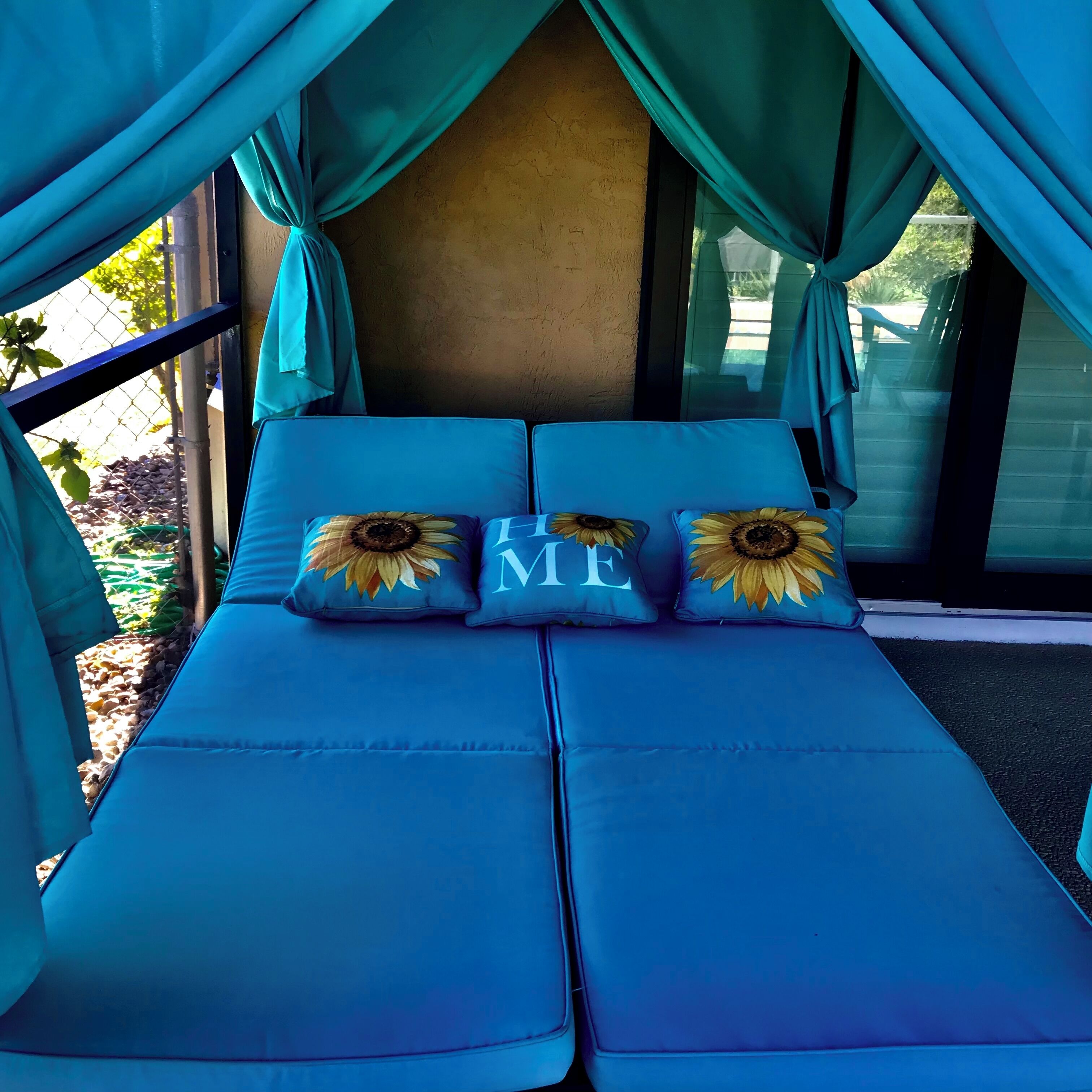 A cozy outdoor seating area with a canopy, two blue cushions with sunflowers, and a wall with a textured surface.