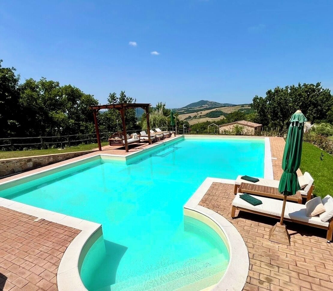 Une piscine rectangulaire dont le centre est d’un bleu clair, entourée d’une terrasse en briques. On y trouve des chaises longues et des parasols.