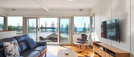 A modern living room with a leather sofa, a small table, a TV, and a balcony with a view of trees and a building.