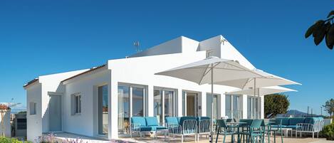 Un edificio moderno blanco con amplios ventanales, mobiliario exterior con cojines azules y un patio.