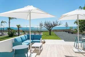 Un patio con un sofá azul, mesas blancas y sombrillas que da a una piscina y una playa.