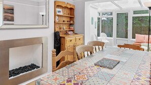 Een ruime kamer met een grote eettafel, houten stoelen, een open haard en een groot raam met uitzicht op de omgeving.