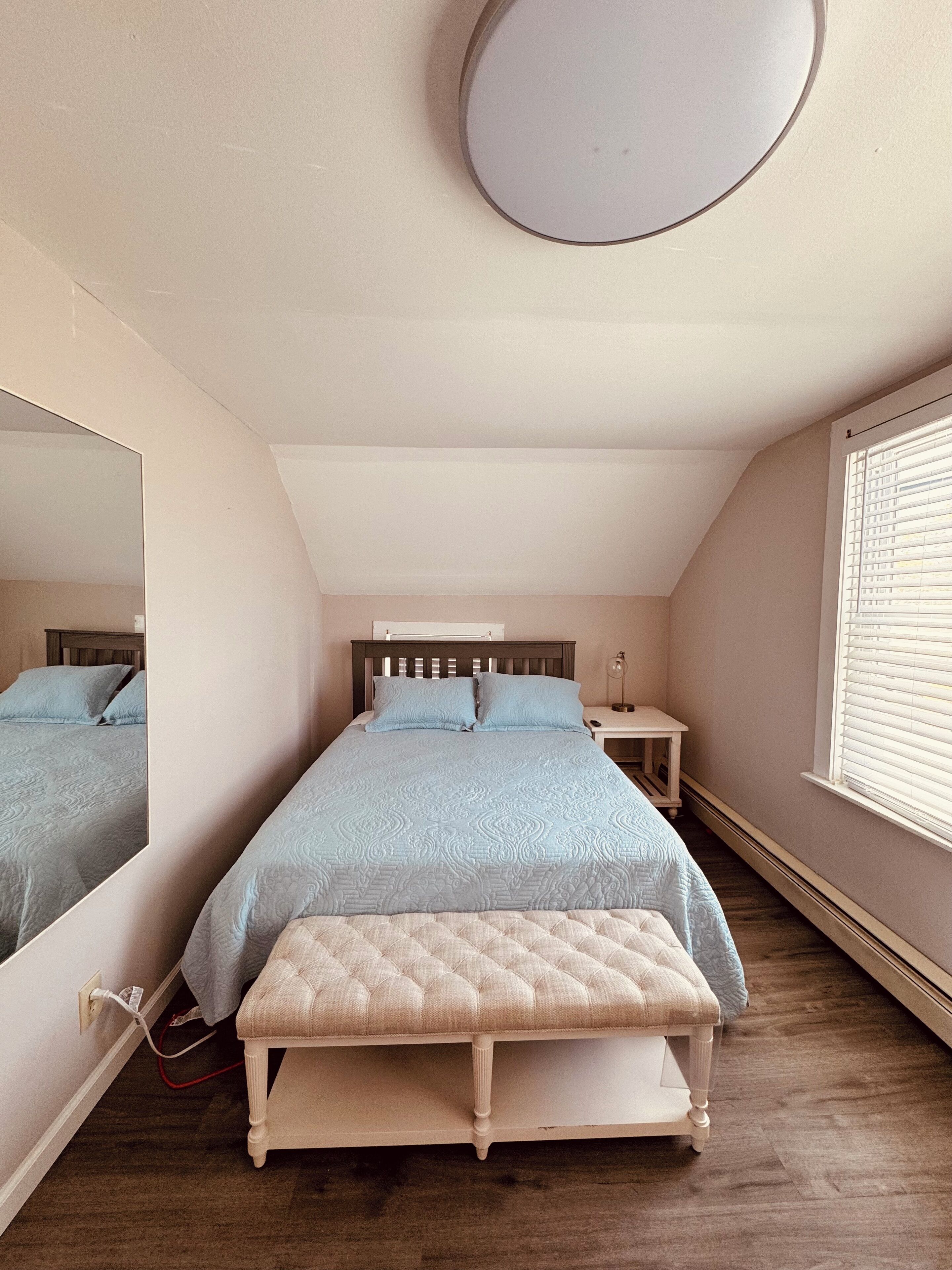 Ein Schlafzimmer mit Bett, Nachttisch, Spiegel und Fenster mit Jalousien.