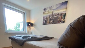 Un dormitorio moderno con una cama, una lámpara de pie, una planta pequeña y un cuadro de un paisaje urbano en la pared.