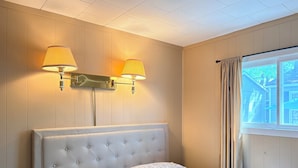Un dormitorio con cabecera capitonada, dos lámparas de noche, una ventana con cortinas y una colcha estampada.