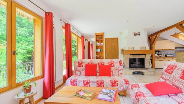 A cozy living room with a sofa, a coffee table, and a fireplace. There are large windows with curtains, a bookshelf, and a kitchen area visible.
