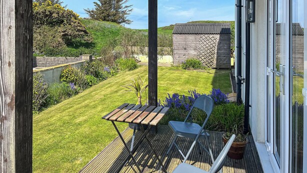 A wooden deck with a table and chairs, overlooking a well-maintained garden with a lawn, various plants, and a wooden structure in the background.