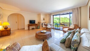 A spacious living room with a sofa, ottoman, and a dining area with chairs and a table. There is a television and a lamp on a side table. - Apartment Santa Monica (Albufeira)