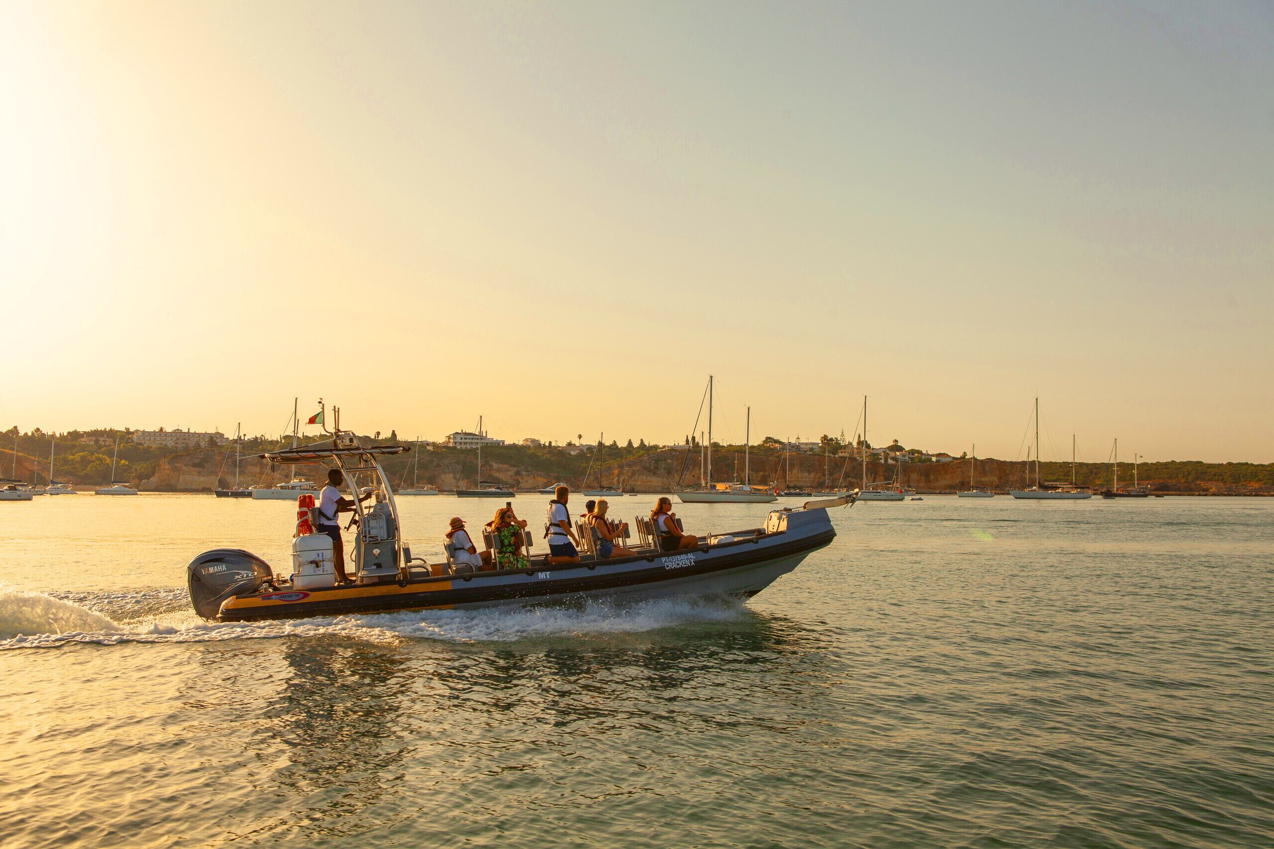 A boat with passengers, moving on a body of water with other boats and a distant shoreline in the background.
