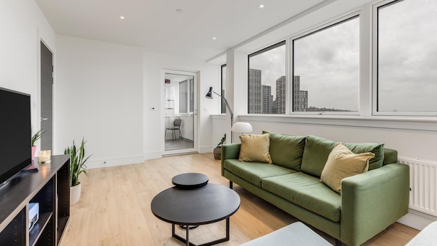 Una sala de estar moderna con un sofá verde, una mesa de centro negra y un televisor sobre un soporte negro. Hay ventanas grandes con vistas a la ciudad y una puerta que conduce a otra habitación.