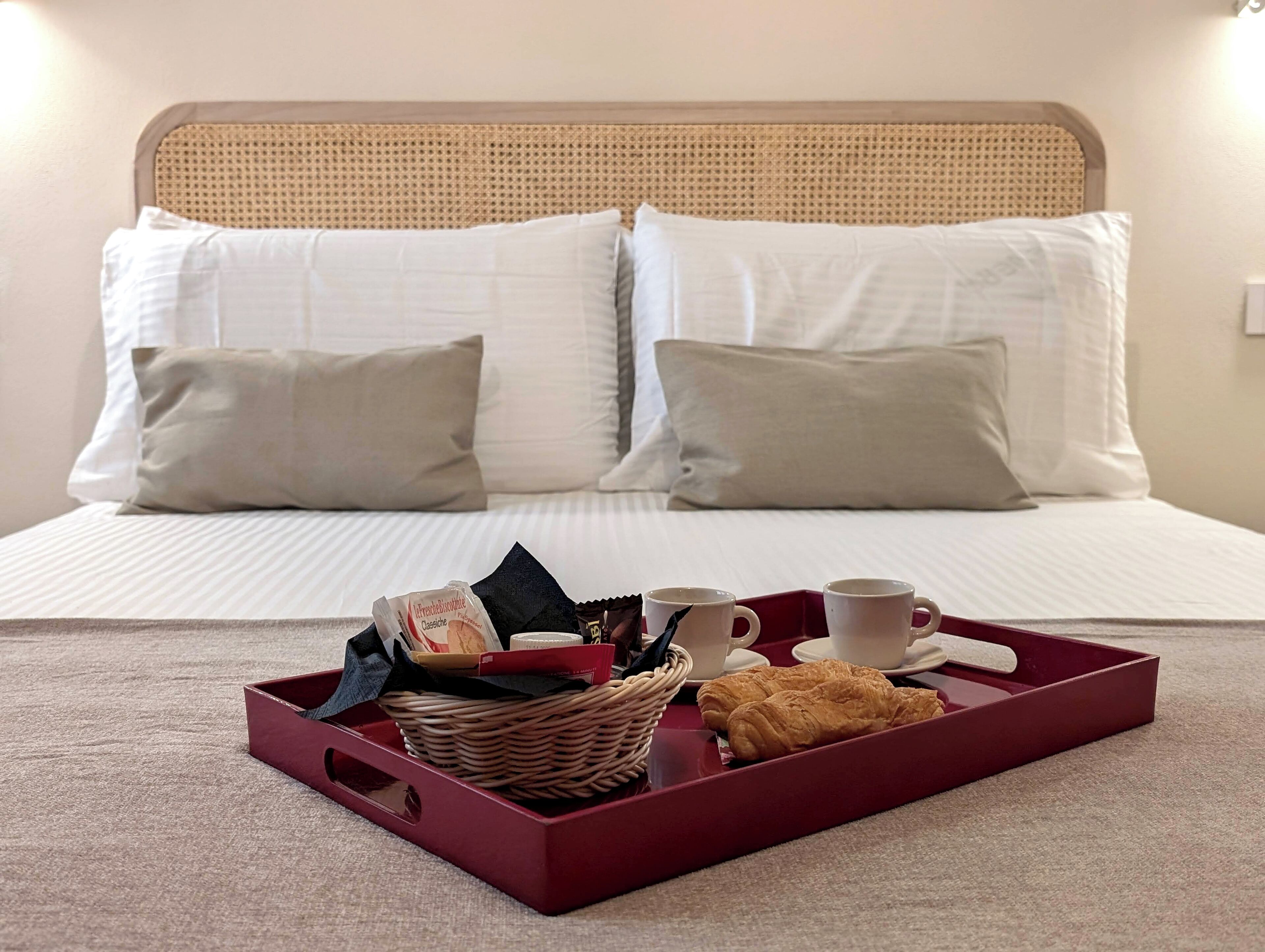A neatly made bed with white linens and two pillows, a tray with croissants, a cup, and a box of chocolate.