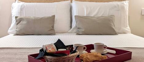 A neatly made bed with white linens and two pillows, a tray with croissants, a cup, and a box of chocolate.