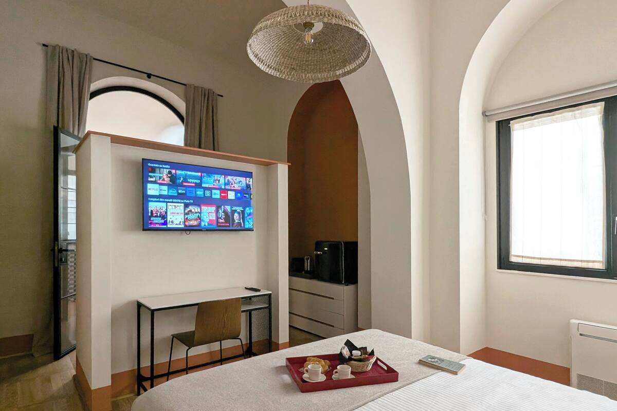Ein Hotelzimmer mit Bett, Schreibtisch, Wandfernseher, Fenster und einer Kristallleuchtre.