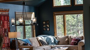 Ein gemütliches Wohnzimmer mit Sofa, Couchtisch und Lampe. Fenster bieten Blick auf Bäume.