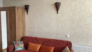 Una habitación con un armario de madera, un sofá rojo con cojines estampados, una mesita redonda y una lámpara de pared.