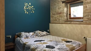 Un dormitorio con cama, mesita de noche, lámpara, estante en la pared y ventana con vistas al exterior.