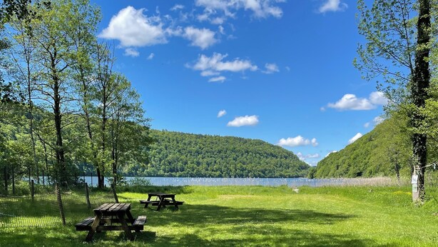 Un área de picnic con una roca grande al frente, un banco de madera y vistas a un lago y colinas boscosas bajo un cielo azul despejado.