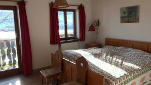 A bedroom with a wooden bed, a desk, and a chair. There are windows with red curtains, a hanging lamp, and a painting on the wall. - Landhaus - Nahe Starnberger See (Eberfing)
