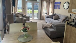 A modern living room with a grey sofa, a dining table with a vase of flowers, and a large window with curtains. - Luxury Rural Hottub Lodge in Northumberland (Morpeth)