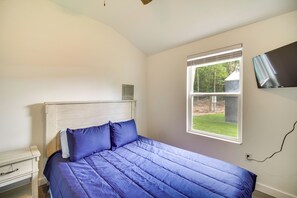 Un dormitorio con cama, mesita de noche, ventana con persianas y televisor montado en la pared.