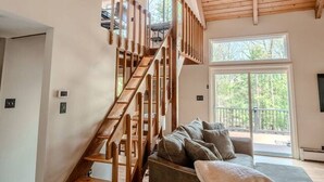 Een ruime kamer met een houten trap, een grijze bank en een groot raam met uitzicht op de omgeving.