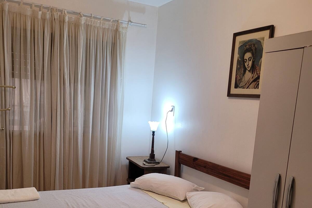 Un lit bien fait, avec un couvre-lit blanc et des oreillers, une lampe de chevet, une tête de lit en bois, une armoire et un tableau encadré au mur.