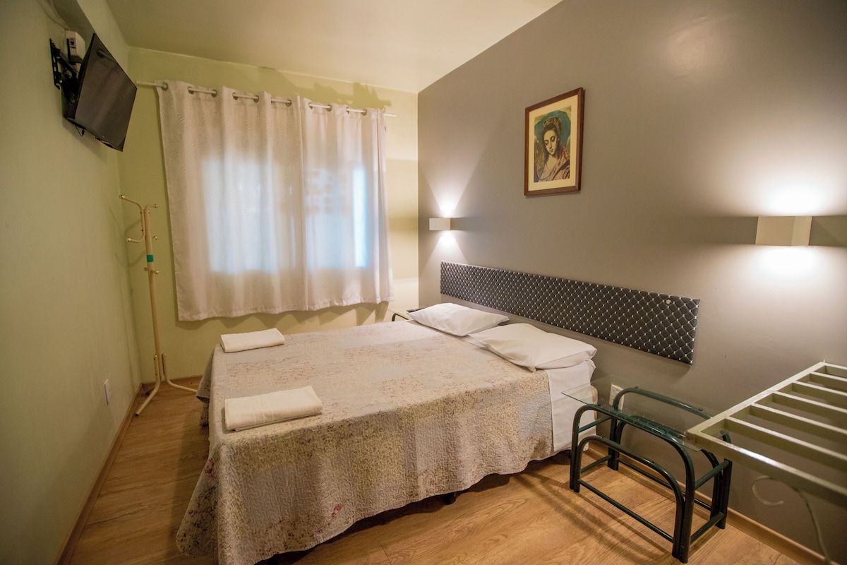 Una camera da letto con un letto, una testiera, una finestra con tende, un quadro appeso al muro e un comodino.