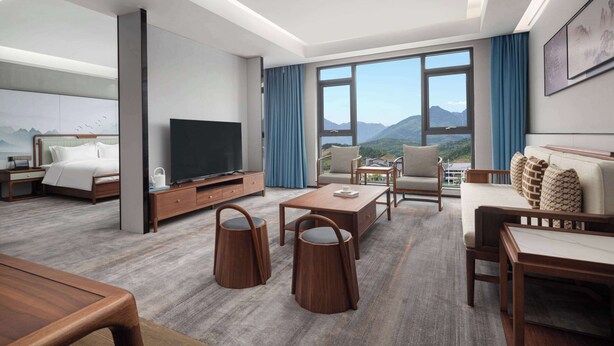 Una habitación de hotel moderna con una cama grande, un televisor de pantalla plana, un sofá, una mesa de centro y vistas a montañas a través de amplias ventanas.