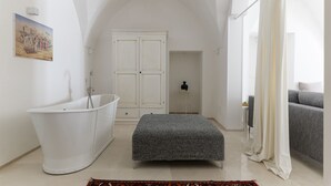 A modern bathroom with a freestanding bathtub, a large arched ceiling, a grey bench, and a grey sofa in the adjacent living area. - Bianca Fortezza by Wonderful Italy (Ostuni)
