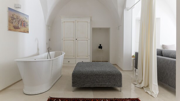 A modern bathroom with a freestanding bathtub, a large arched ceiling, a grey bench, and a grey sofa in the adjacent living area. - Bianca Fortezza by Wonderful Italy (Ostuni)