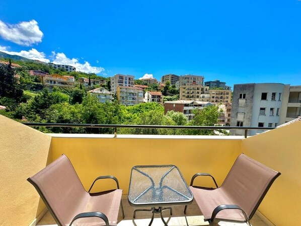 Un balcón con dos sillas y una mesa, con vistas a un paisaje urbano con edificios y áreas verdes.