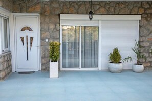 Fachada de piedra con una puerta blanca y una puerta corrediza de vidrio. Hay tres macetas con plantas frente a la puerta corrediza.