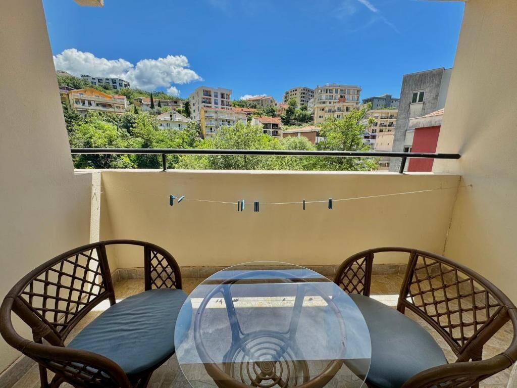 A balcony with a glass table and two wicker chairs, overlooking a residential area with buildings and greenery.