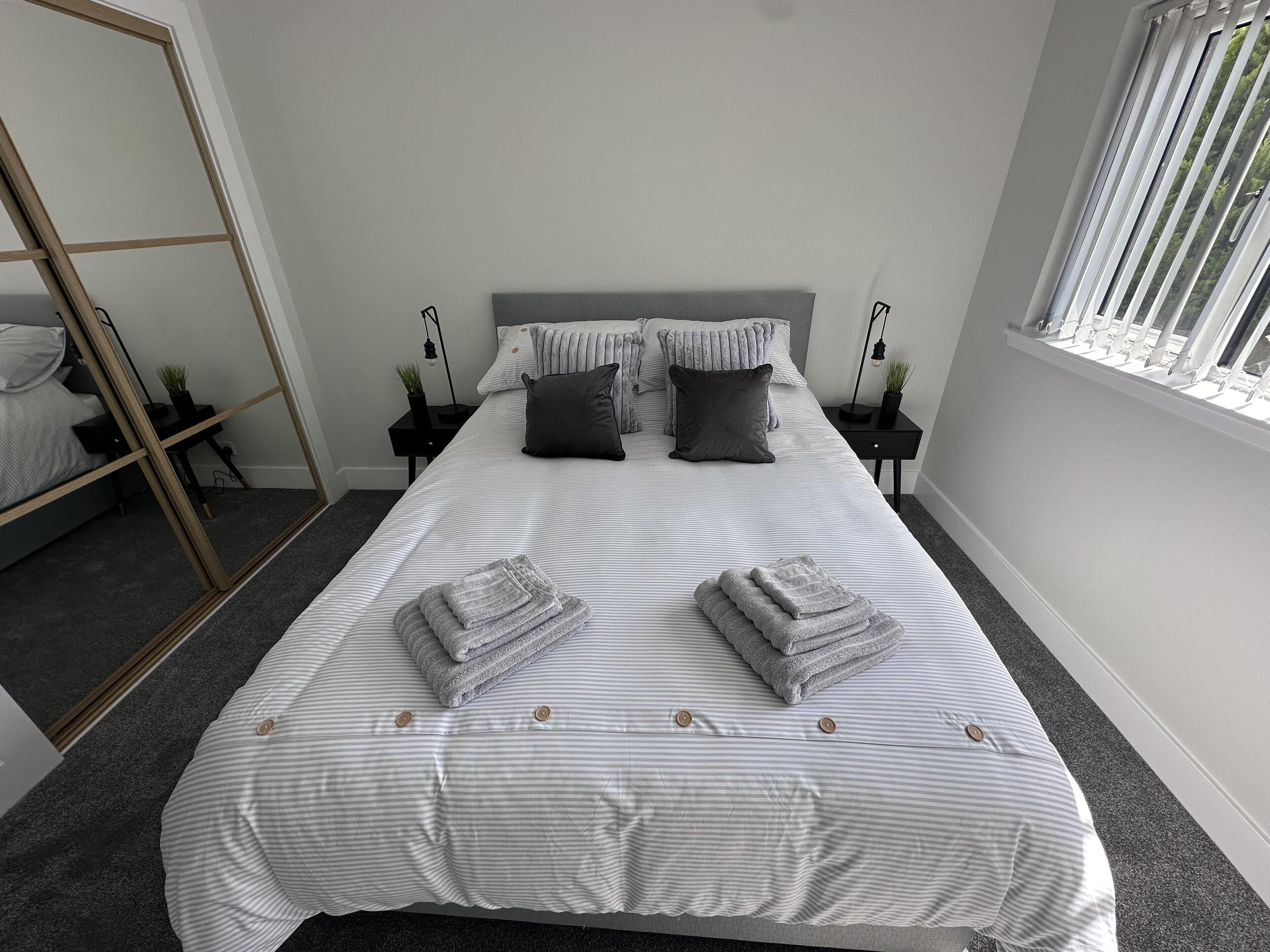 Una cama bien hecha con ropa de cama a rayas, dos almohadas oscuras y toallas dobladas. Hay mesitas de noche con lámparas y un espejo.