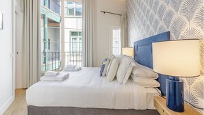 Een hotelkamer met een bed, een nachtkastje met een lamp, en uitzicht op een balkon met leuningwerken.