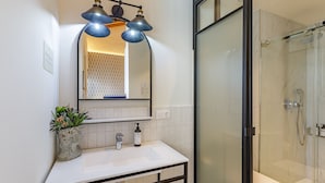 A modern bathroom with a white sink, a mirror with three pendant lights, a glass shower enclosure, and a potted plant on the sink. - Palacio Vegafuente 1.3 (Málaga)