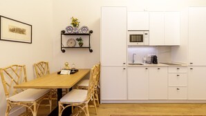 Een eetgedeelte met een houten tafel en rieten stoelen, een klein schap met decoratieve borden, en een keuken met witte kasten en een magnetron.