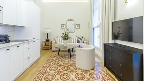 Een moderne woonkamer met een bank, salontafel, poef, een televisie en een keukenhoek met kastjes en een koffieapparaat.
