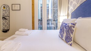 Hotelkamer met een bed, witte lakens, een spiegel met bladmotief, een raam met uitzicht op een balkon, en een kussen met blauw-wit patroon.